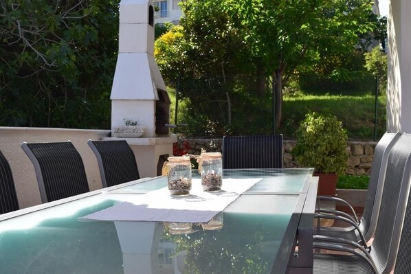 An outdoor dining area with a glass table, metal chairs, and a white barbecue grill in the background. - Apartment in Banjol with Balcony, Air condition, WIFI, Dishwasher (4888-1) (Banjol)