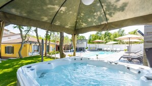 Un jacuzzi bajo una pérgola, con una piscina y sillones de descanso al fondo.