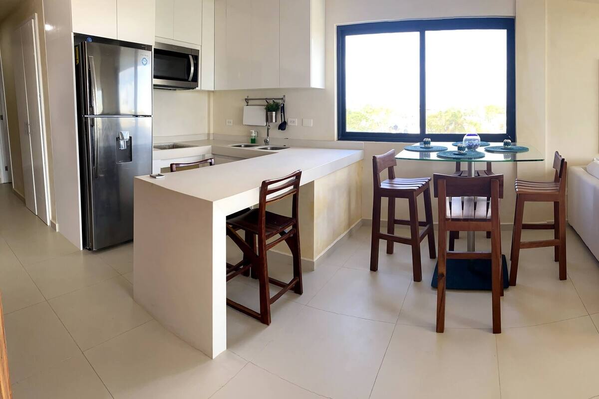 Eine moderne Küche mit Kühlschrank, Mikrowelle und Essbereich mit Stühlen und Tisch. Im Wohnbereich befindet sich eine weiße Couch und ein Fernseher.