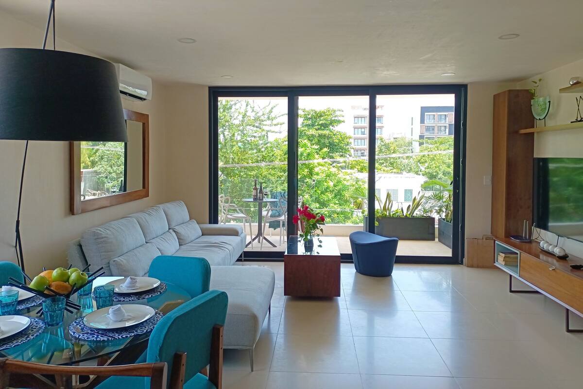 Ein modernes Wohnzimmer mit einer Couch, Esstisch und Stühlen, einem Balkon mit Blick auf Grünflächen und Gebäude sowie einem Flachbildfernseher auf einem Holzboden.