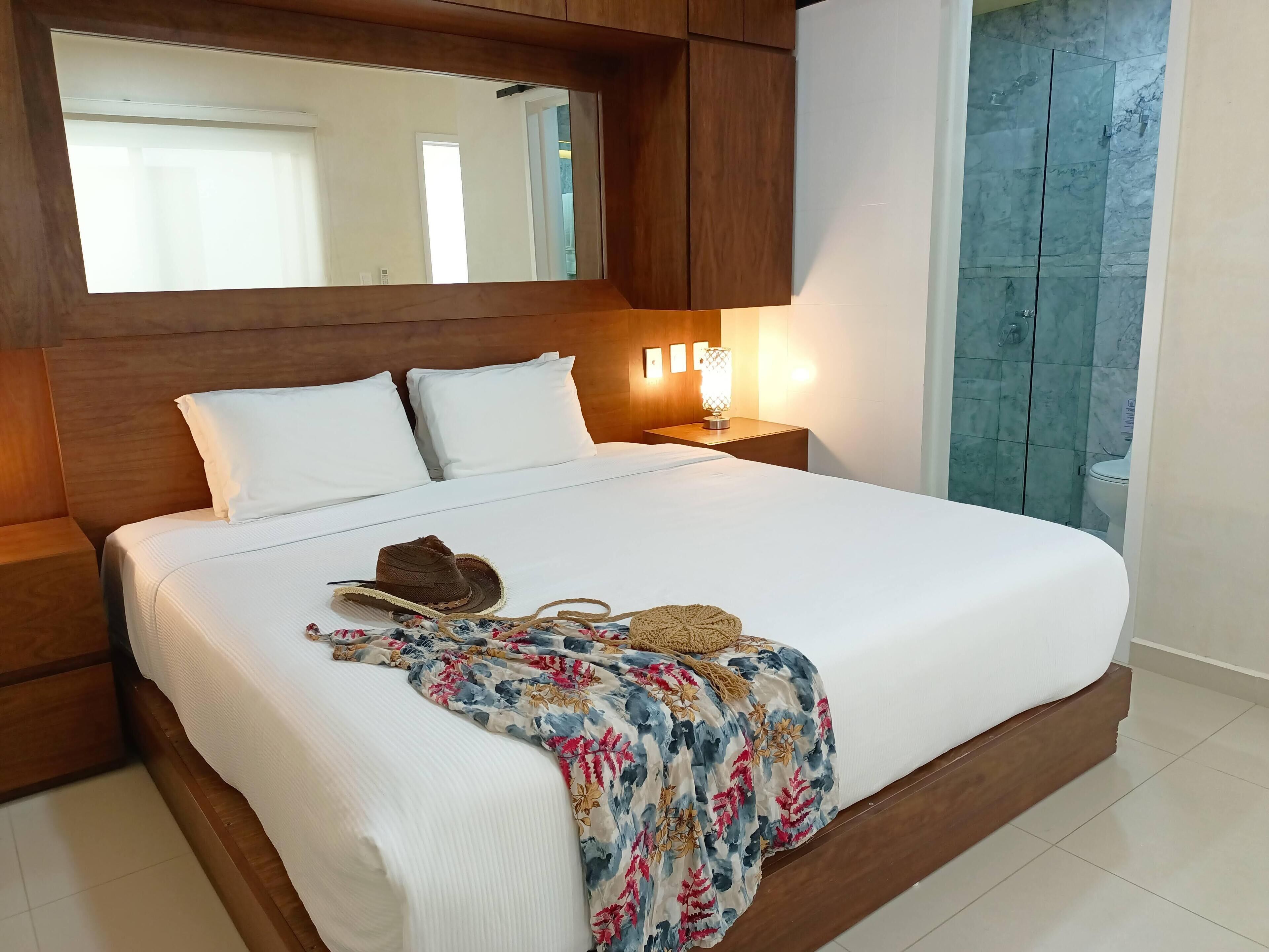 A neatly made bed with white linens, a floral blanket, and a hat on the bed. There is a nightstand with a lamp and a mirror above the bed. The room has wooden headboard and nightstands, and a bathroom with a glass shower door is visible.