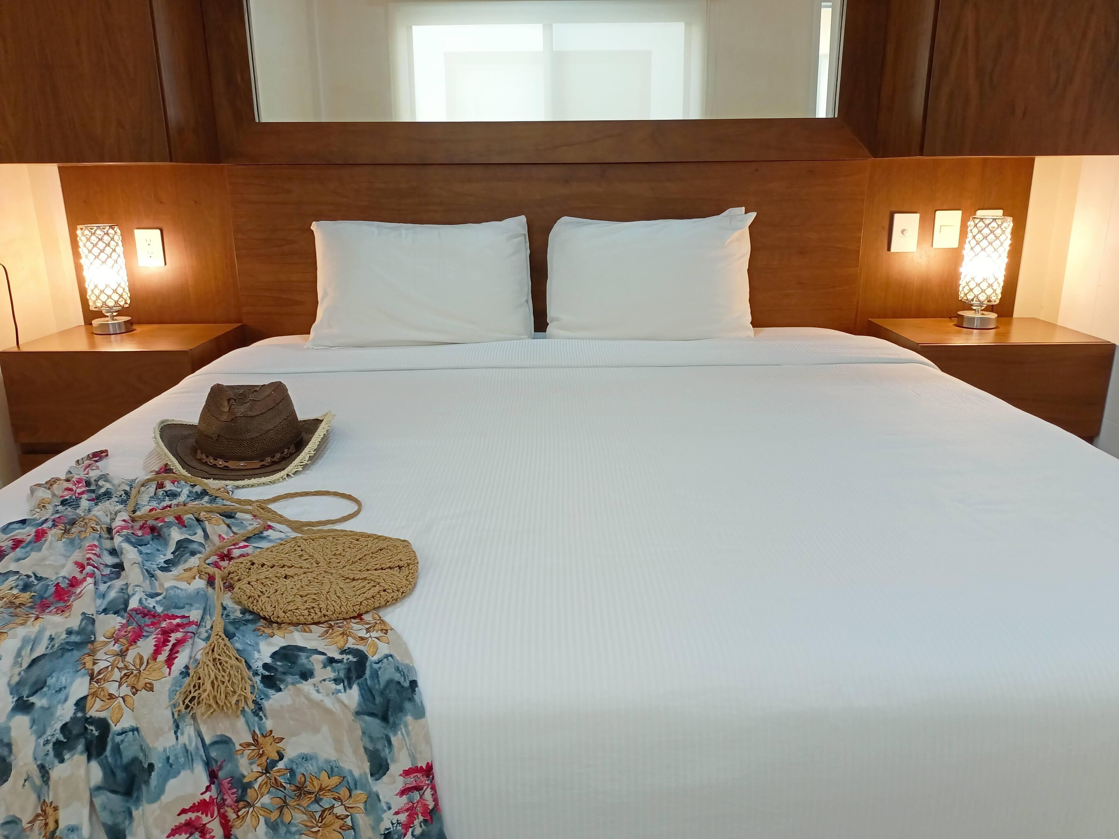 Un lit bien fait, avec des draps blancs, une couverture à motifs floraux, un chapeau tressé et un petit sac tressé posés sur le lit.