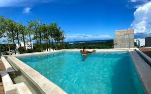 Una piscina infinita rectangular con agua azul cristalina, rodeada por una terraza de madera y un muro de piedra.