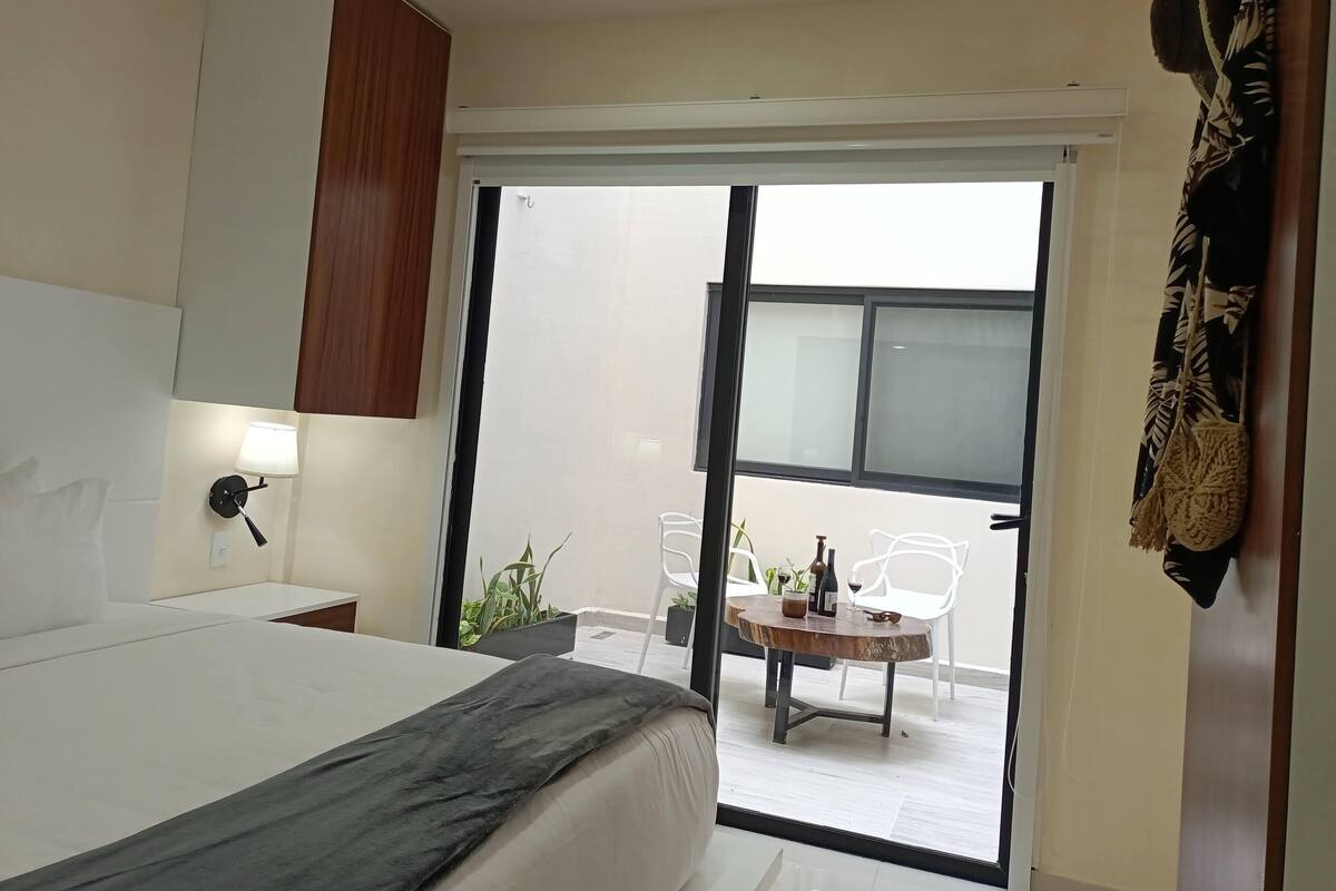 Ein Schlafzimmer mit Bett, Nachttisch, Lampe, Kleiderschrank und einer Glastür, die auf einen Balkon mit Tisch, Stühlen und Pflanzen führt.