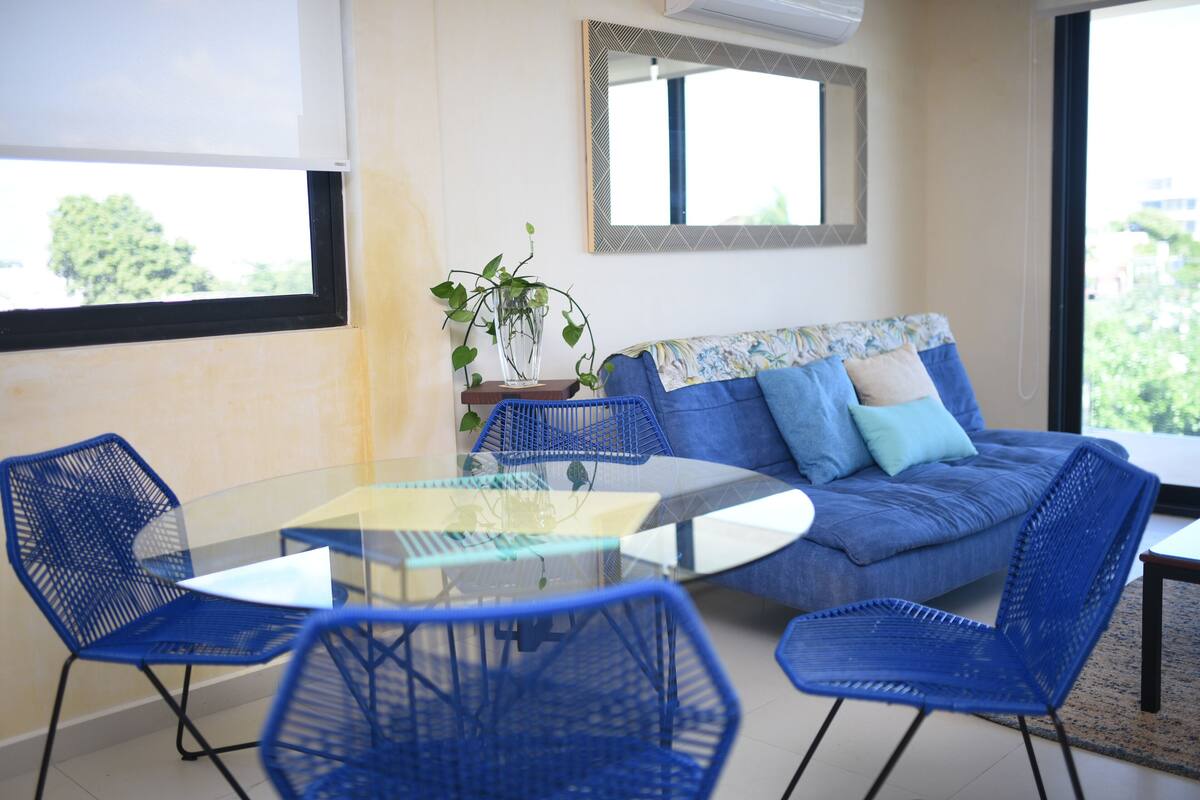 Ein modernes Wohnzimmer mit einem Glastisch, blauen Sesseln, einer Couch mit gemusterten Kissen und einem großen Spiegel an der Wand.