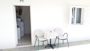 En liten sittplats utomhus med ett runt bord och två stolar, en dörr som leder in till ett rum med en mikrovågsugn och ett fönster med gardiner.