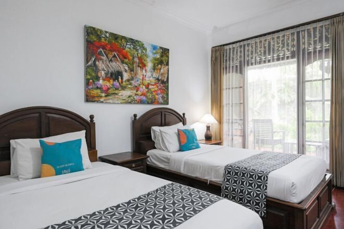 A hotel room with two beds, a large window with curtains, a painting on the wall, and a nightstand with a lamp.
