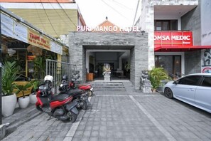 Un edificio con un cartello che riporta 'Puri Mango Hotel' e 'OMSAS MEDIC' sulla facciata. Sono presenti piante in vaso e un'auto parcheggiata di fronte.