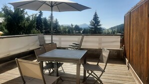Una terraza de madera con una mesa, sillas, un paraguas y vistas a árboles y montañas.