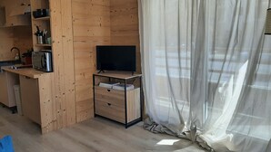 Un espacio habitable compacto con kitchenette, televisor de pantalla plana y un amplio ventanal con cortinas translúcidas.