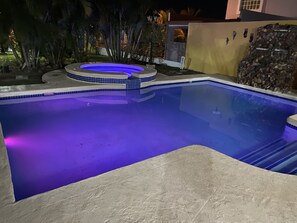 A swimming pool with a blue-lit hot tub area, surrounded by a stone wall and palm trees.
