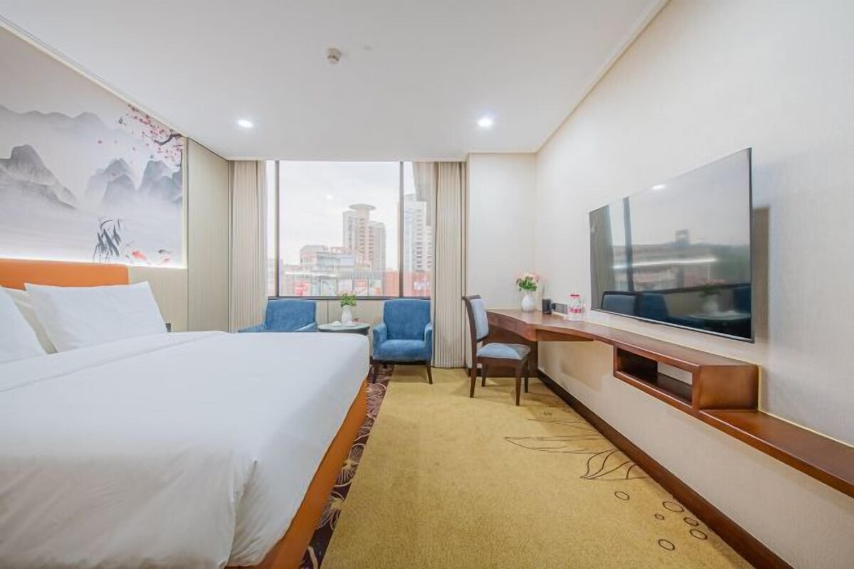 A hotel room with a bed, a desk, a chair, a TV, and a view of the cityscape.