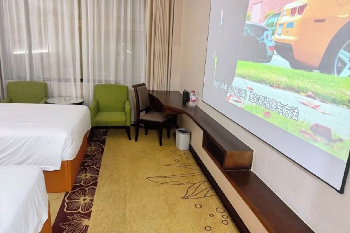 A hotel room with two beds, a green armchair, a small table, and a TV screen displaying an image of a car.