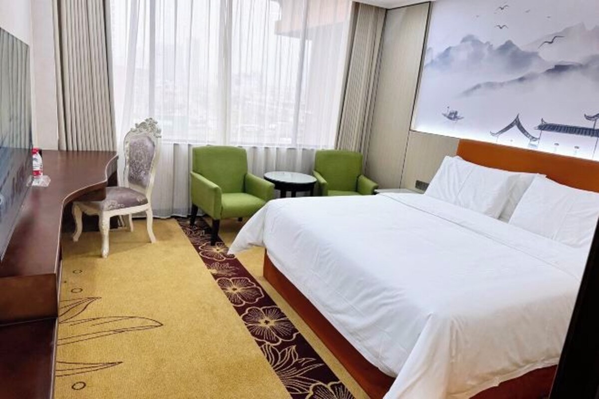 A hotel room with a large bed, two green armchairs, a small round table, and a chair with a cushion.