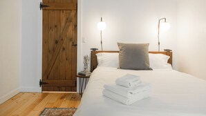 Un dormitorio con puerta de madera, una cama con sábanas blancas, dos lámparas de noche y una mesita de noche con un libro y un florero.