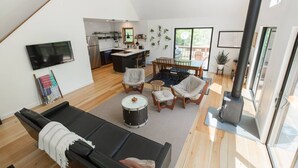 Un amplio espacio de sala con un sofá negro, un set de batería y una mesa de comedor con sillas. Se ve una cocina al fondo, con un gran ventanal.