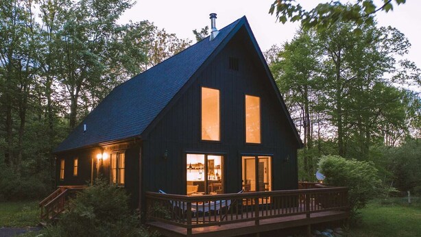 Una cabaña de madera oscura con techo inclinado, ventanas grandes y una terraza de madera.