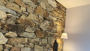 A bed with dark bedding, a colorful pillow, a stone wall, and a nightstand with a lamp. - Longere LE Penty - Plougonvelin (Plougonvelin)