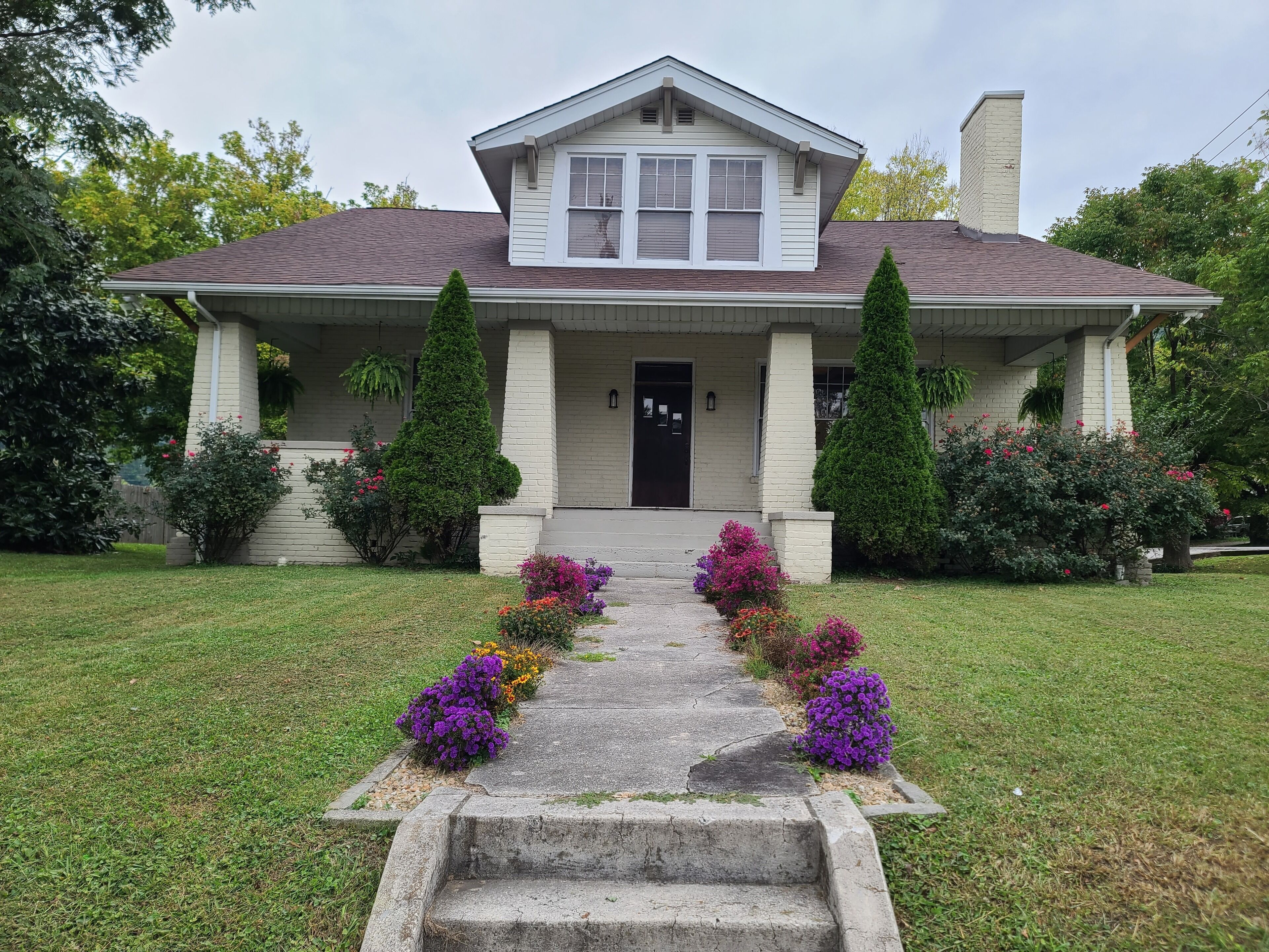 A two-story house with a front porch, a chimney, and a well-maintained lawn with colorful flowers.