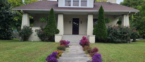 A two-story house with a front porch, a chimney, and a well-maintained lawn with colorful flowers.