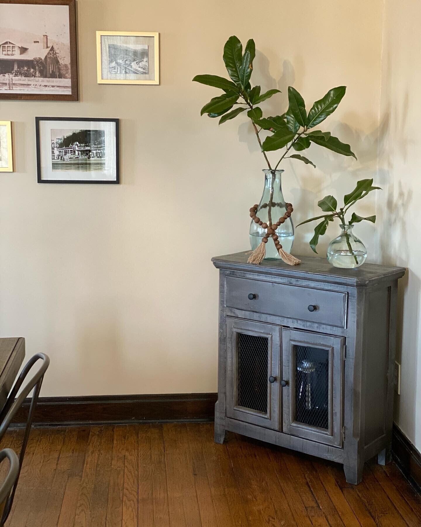 A corner with a small wooden cabinet with two doors, a plant, and two framed pictures on the wall.