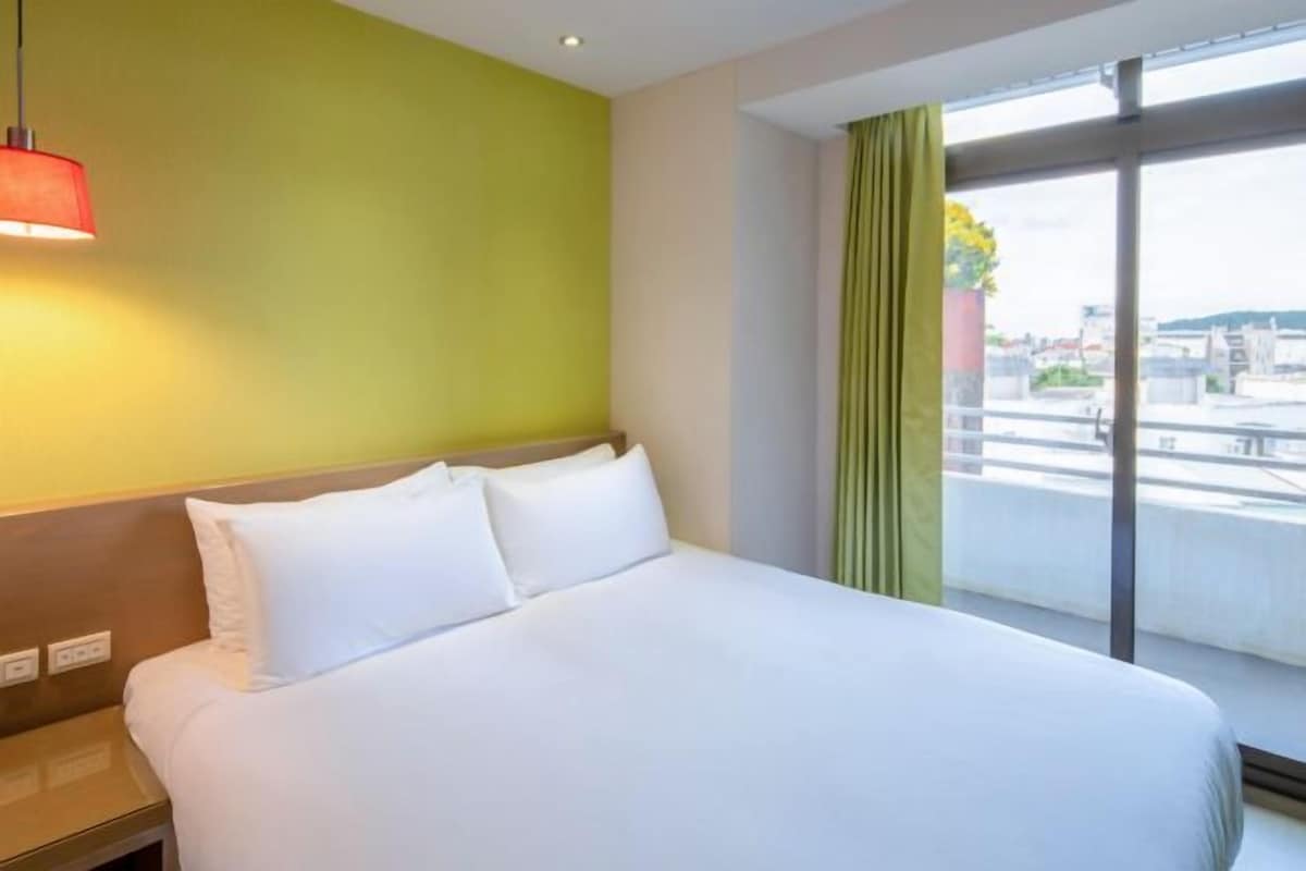 A hotel room with a bed, a nightstand, a red pendant light, a green wall, a window with curtains, and a view of the cityscape.