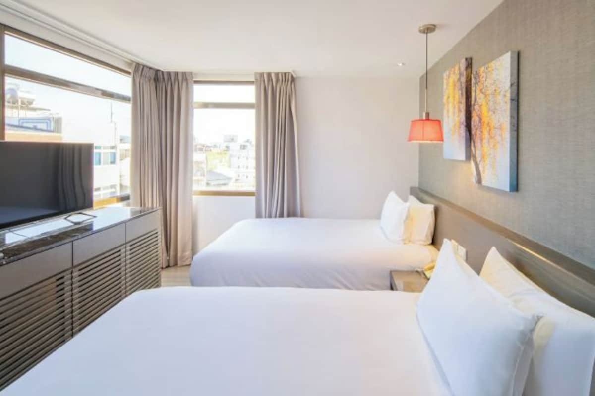 A hotel room with two beds, a TV on a dresser, and a large window with curtains.