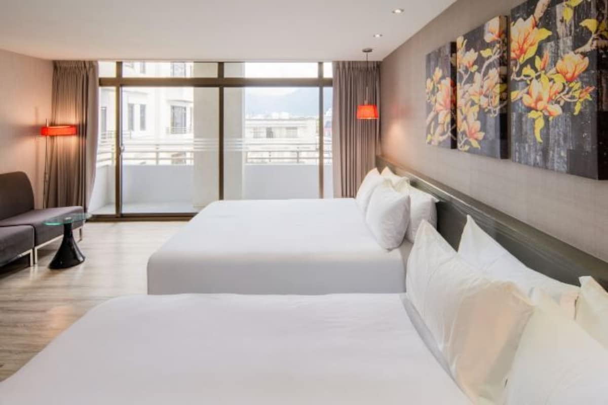 A hotel room with two beds, a sofa, a glass table, and a view of the city through large windows.