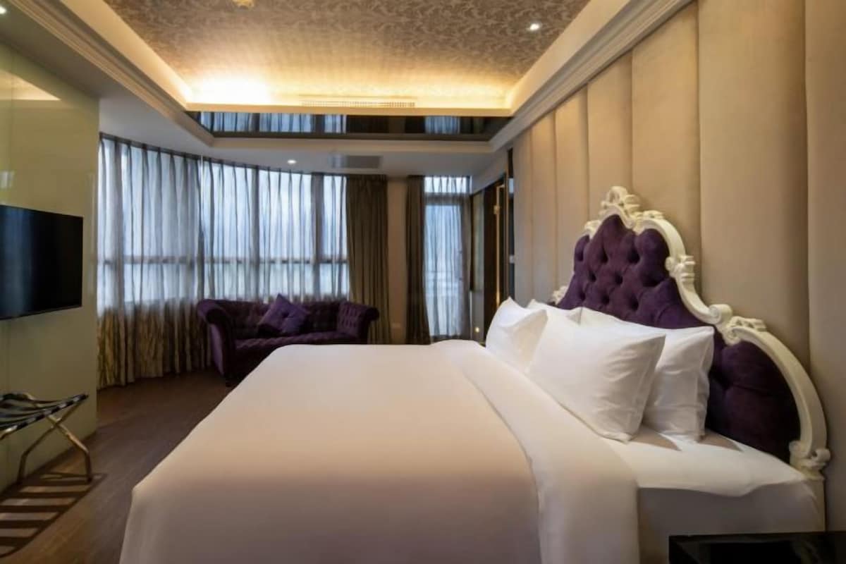 A hotel room with a large bed, a TV, a sofa, and a view of the city through large windows.