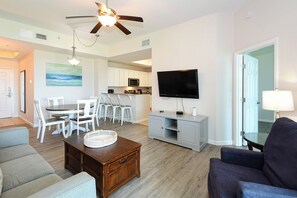 A living room with a ceiling fan, a flat-screen TV, a dining area with white chairs, and a kitchen in the background.