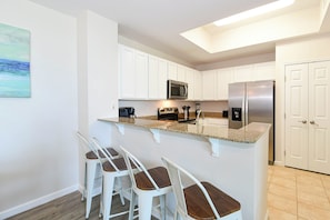 A kitchen with white cabinets, a granite countertop, a stainless steel refrigerator, and a microwave. There are three white bar stools with wooden seats. - 1st Floor condo at The Palms of Destin with Free Beach Shuttle! (Destin)