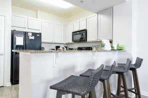 A kitchen with white cabinets, a black refrigerator, a microwave, and a bar area with wicker stools.