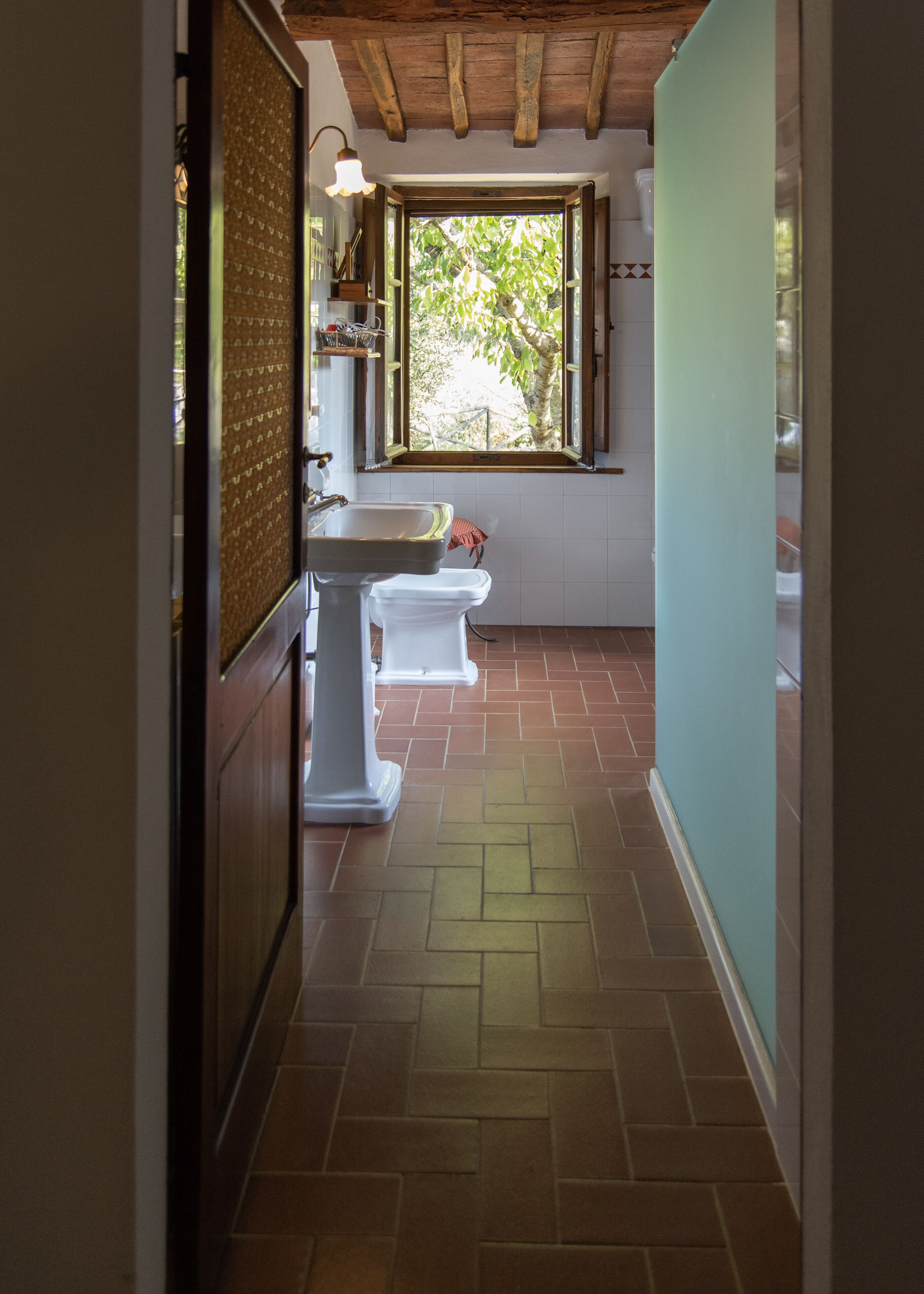 Un couloir étroit, dont le sol est recouvert de carreaux, mène à une salle de bain équipée d’une fenêtre, d’un lavabo et de deux toilettes.