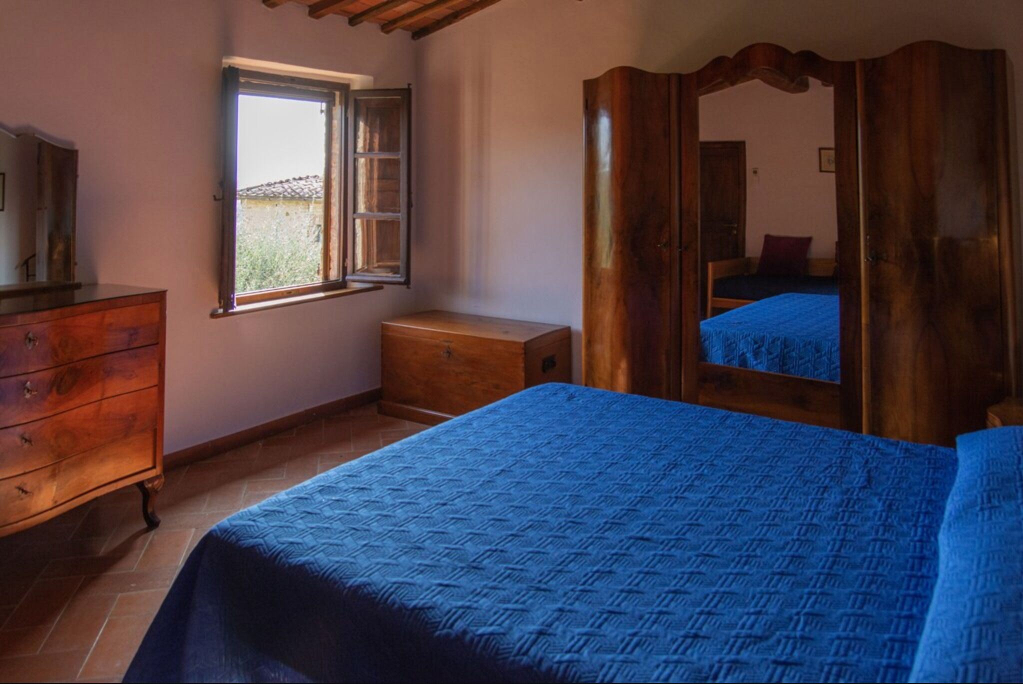 A bedroom with a wooden dresser, a small bedside table, a bed with a blue bedspread, a window with an open shutter, and a wooden wardrobe with a mirror.