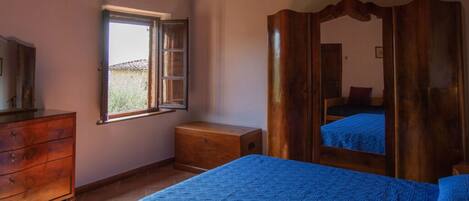 A bedroom with a wooden dresser, a small bedside table, a bed with a blue bedspread, a window with an open shutter, and a wooden wardrobe with a mirror.