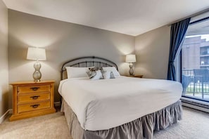 A bedroom with a bed, two bedside tables with lamps, a window with curtains, and a view of buildings outside. - Lake Dillon! Ampitheatre, Walk to Lake,Pool&HT (Dillon)