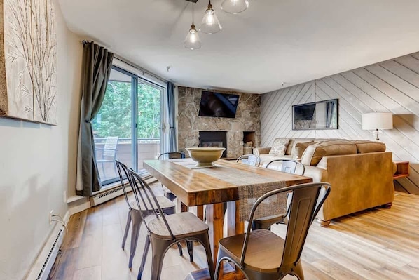 A modern living room with a fireplace, a dining area with a wooden table and chairs, and a large window with curtains. - Lake Dillon! Ampitheatre, Walk to Lake,Pool&HT (Dillon)