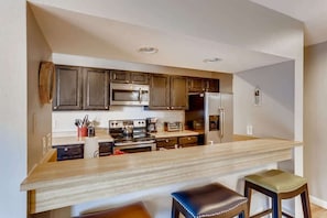 A modern kitchen with dark wooden cabinets, stainless steel appliances, a wooden countertop, and two stools. - Lake Dillon! Ampitheatre, Walk to Lake,Pool&HT (Dillon)