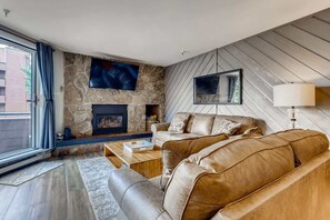 A modern living room with a stone fireplace, leather sofa, and a large TV mounted on the wall. - Lake Dillon! Ampitheatre, Walk to Lake,Pool&HT (Dillon)
