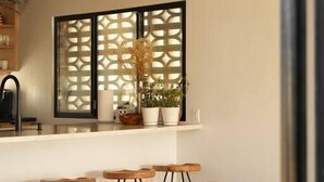 Un salon moderne avec un canapé en cuir marron, un coin cuisine doté d’un plan de travail blanc et une fenêtre décorative ornée d’un motif en treillis.
