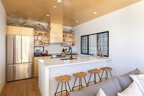 A modern kitchen with a stainless steel refrigerator, wooden bar stools, and a large window with a geometric pattern. - Neutra Home Joshua Tree Luxury Retreat Pool and Spa (Joshua Tree)