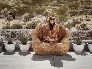 A person seated on a woven chair outdoors with potted plants around. - Neutra Home Joshua Tree Luxury Retreat Pool and Spa (Joshua Tree)