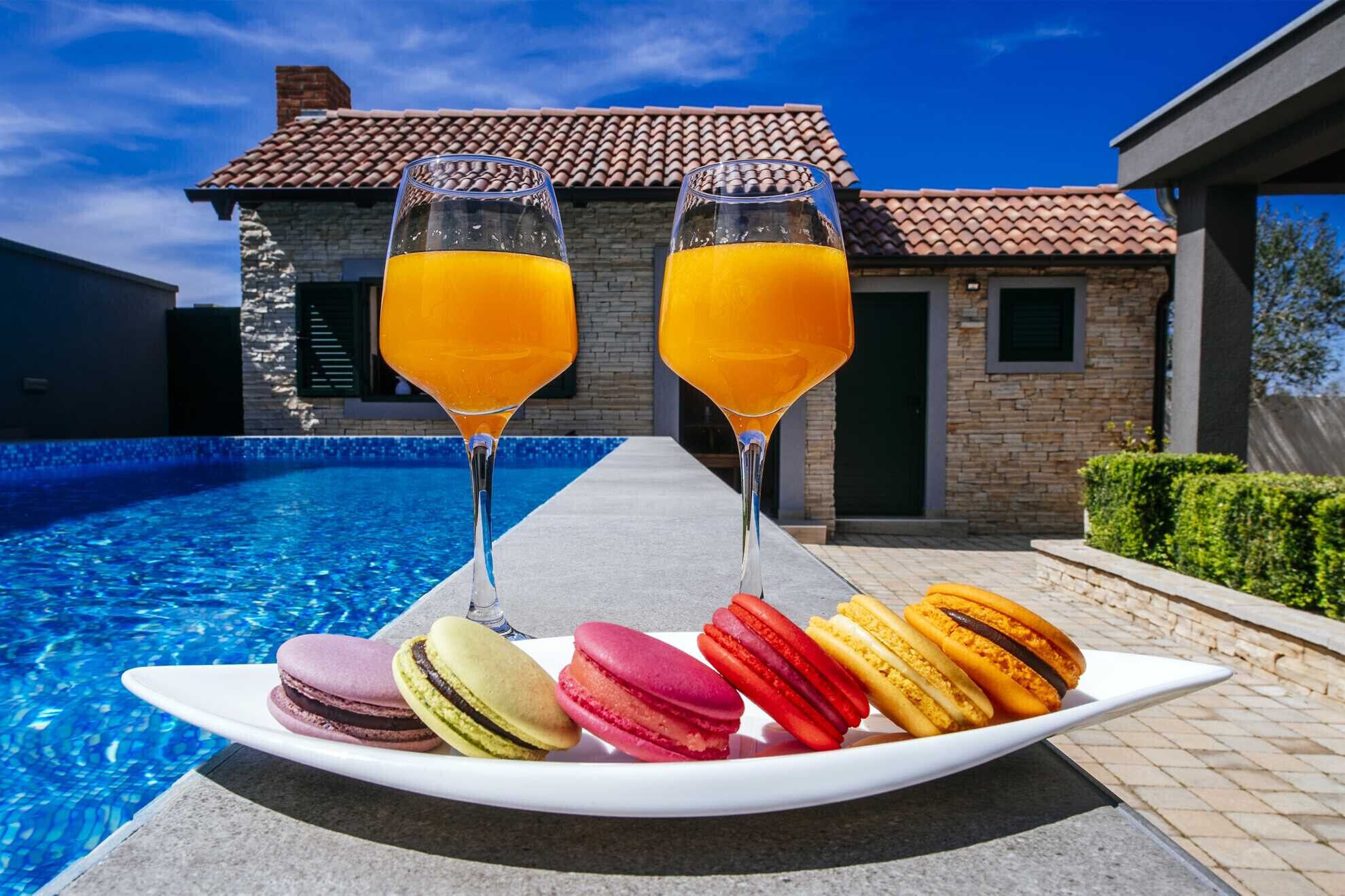 A poolside setting with two glasses of orange juice, a plate of colorful macarons, and a house with a tiled roof in the background.
