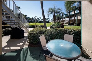 A patio area with a round table and chairs, surrounded by greenery and a view of a building with a staircase.
