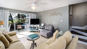 A living room with beige sofas, a grey armchair, a TV, and a decorative wall piece. - Seaspray Condos - 100B (Fort Walton Beach)