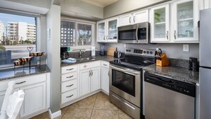 A modern kitchen with white cabinetry, stainless steel appliances, and a large window offering a view of the city. - Seaspray Condos - 100B (Fort Walton Beach)