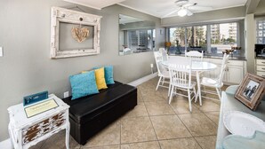 A modern living room with a black sofa, white chairs, a dining table, and a kitchen area visible through large windows. - Seaspray Condos - 100B (Fort Walton Beach)