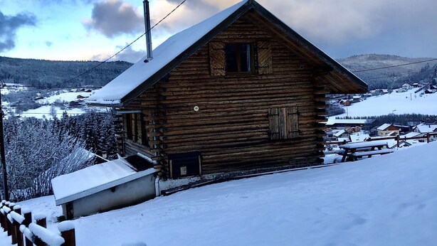 Een houten hut met een sneeuwbedekte dak, omgeven door een besneeerd landschap en verre bergen.