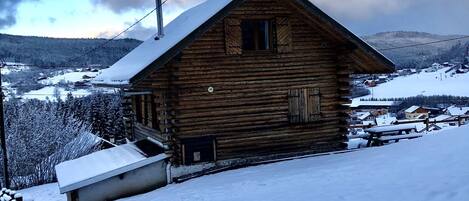 Une cabane en bois, dont le toit est recouvert de neige, entourée d’un paysage enneigé et de montagnes lointaines.
