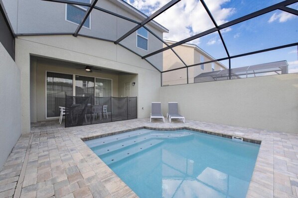 Piscina com estrutura de vidro, duas espreguiçadeiras e área de estar com mesa e cadeiras.